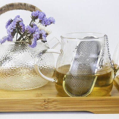 Boule à Thé ovale en maille d'acier dans une tasse en verre avec une théière décorée de fleurs mauves en arrière-plan