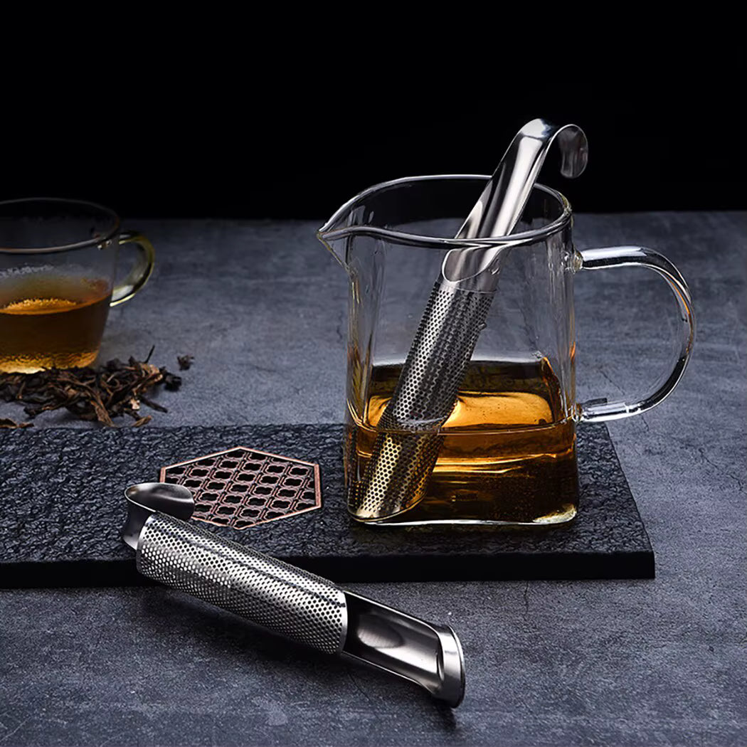 Composition sophistiquée de la Cuillère à Thé | Élégance Inox suspendue dans une carafe en verre remplie de thé, et un autre infuseur ouvert posé à cöté sur un dessous de verre en marbre noir