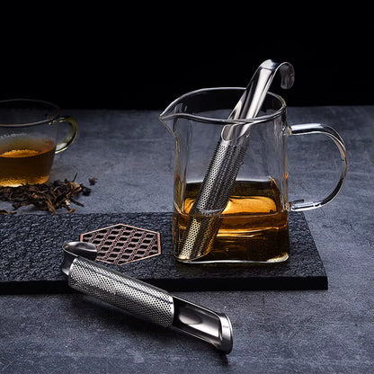 Composition sophistiquée de la Cuillère à Thé | Élégance Inox suspendue dans une carafe en verre remplie de thé, et un autre infuseur ouvert posé à cöté sur un dessous de verre en marbre noir