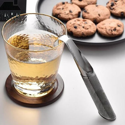 Cuillère à thé | Élégance Inox - suspendue au bord d'un verre d'eau chaude martelé. Une assiette de cookies aux pépites de chocolat est visible en arrière-plan