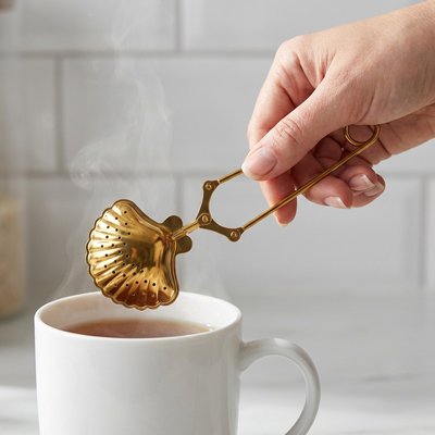 Gros plan sur l' Infuseur a thé Pince-Moi en forme de coquillage doré au-dessus d'une tasse de thé chaude.