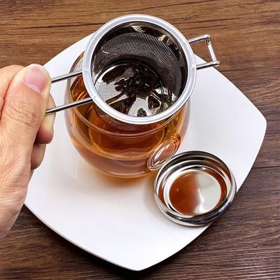 Infuseur à thé en inox 304 avec doubles poignées infusant dans un verre transparent avec sa coupelle de récupération
