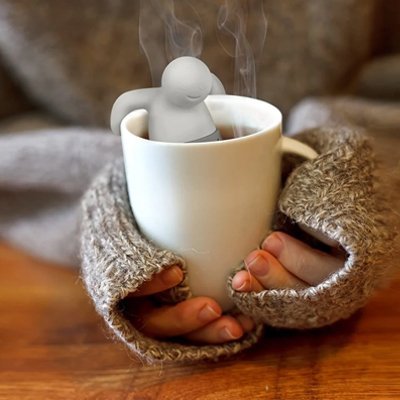 Infuseur à thé | Petit Bonhomme - accroché sur le bord d'une tasse blanche entouré par des mains