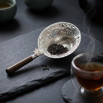 Passoire à thé Inoxia argent posée sur planche ardoise noire près d'une tasse de thé en verre