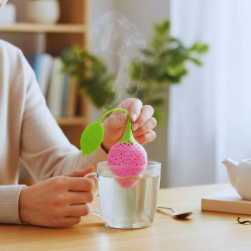 Plan rapproché d'une main abaissant un Infuseur a Thé | Fraisinette rose dans l'eau chaude d'une tasse en verre