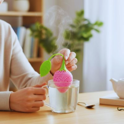 Plan rapproché d'une main abaissant un Infuseur a Thé | Fraisinette rose dans l'eau chaude d'une tasse en verre
