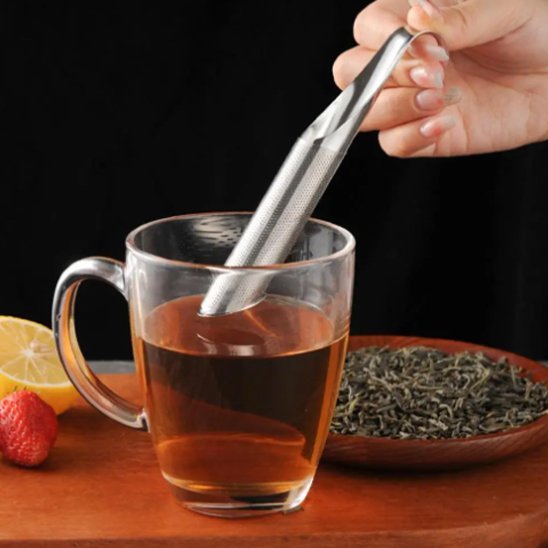 Une main tient la Cuillère à thé | Élégance Inox - en acier biseauté au-dessus d'une tasse transparente avec une assiette de thé en vrac et une fraise sur la table.