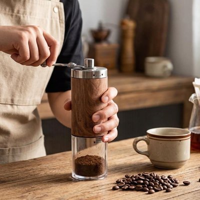 Une personne en tablier moud du café avec le Moulin à Café | Arômes Intenses manuel, posé sur une table en bois rustique à côté d'une tasse de café.